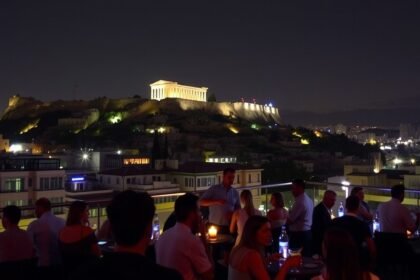 Éjszakai panoráma Athénban az Akropolisz mellett, tetőteraszon ülő vendégekkel.